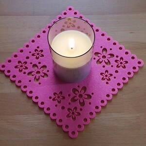 Könnte beinhalten: Eine weiße Kerze in einem Glasgefäß steht auf einem rosa Filzuntersetzer mit Blumenmuster. Die Kerze ist angezündet und die Flamme ist sichtbar.