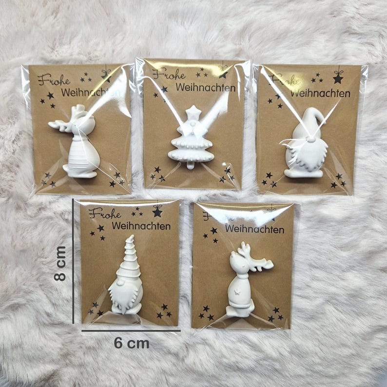May include: Five white Christmas ornaments in clear plastic packaging, each mounted on a brown card. The ornaments include a reindeer, Christmas tree, gnome, and other festive shapes. The cards measure 8 cm high and 6 cm wide, with the text "Frohe Weihnachten".