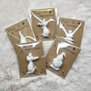 May include: Five small, white Christmas ornaments in clear plastic packaging, each attached to a brown card. The cards have the words "Frohe Weihnachten" and star graphics. The ornaments include a reindeer, gnome, and Christmas tree.