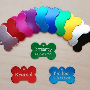 Könnte beinhalten: Sortiment an bunten, knochenförmigen Hundemarken mit eingraviertem Text. Die Marken sind aus Metall und haben ein Loch zum Anbringen an einem Halsband. Der Text auf den Marken lautet "Smarty", "Krümel" und "I'm lost".
