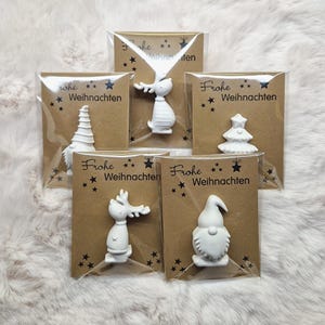 May include: A collection of white Christmas ornaments in various shapes, including a reindeer, gnome, and Christmas tree, each packaged on a brown card with the text "Frohe Weihnachten" and star accents.