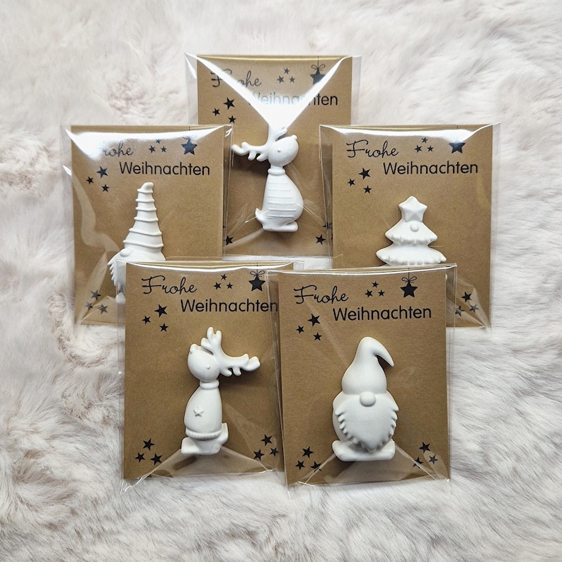 May include: A collection of white Christmas ornaments in various shapes, including a reindeer, gnome, and Christmas tree, each packaged on a brown card with the text "Frohe Weihnachten" and star accents.