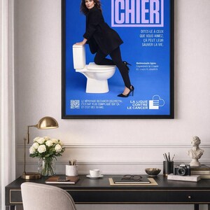 Puede incluir: Un p&oacute;ster enmarcado con fondo azul muestra a una mujer vestida de negro apoyada en un inodoro blanco. El texto "VA CHIER" est&aacute; en letras grandes. Tambi&eacute;n hay texto adicional en franc&eacute;s.