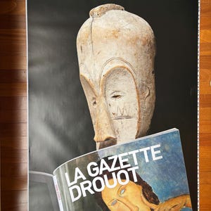 May include: A black and white poster with the text "L'HEBDOMADAIRE DES ARTS ET DES ENCHERES 3,50€" at the top. A wooden sculpture of a face is partially visible in the upper right corner. A magazine with the text "LA GAZETTE DROUOT" is partially visible in the lower right corner.