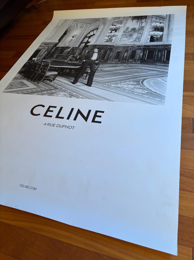 May include: Monochrome poster advertising CELINE, featuring a fashion model in a formal setting. The poster includes the brand name "CELINE", the address "4 RUE DUPHOT", and the website "CELINE.COM". The image is set against a backdrop of a grand interior with a person.
