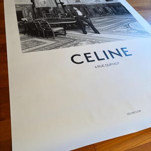 May include: Monochrome poster with a fashion image of a person in a suit, posed near a table in a large room. The word "CELINE" is printed in bold letters, with "4 RUE DUPHOT" beneath it. The website address is also visible.