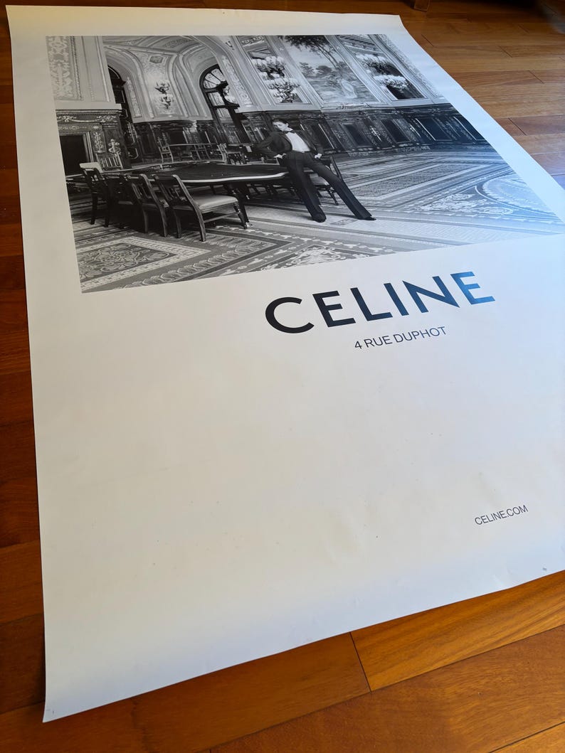 May include: Monochrome poster with a fashion image of a person in a suit, posed near a table in a large room. The word "CELINE" is printed in bold letters, with "4 RUE DUPHOT" beneath it. The website address is also visible.