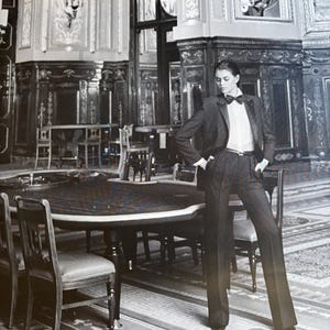 May include: A person in a dark grey suit, white shirt, and black bow tie poses in a room with a large round table and chairs. The room features ornate wall panelling and a patterned rug.