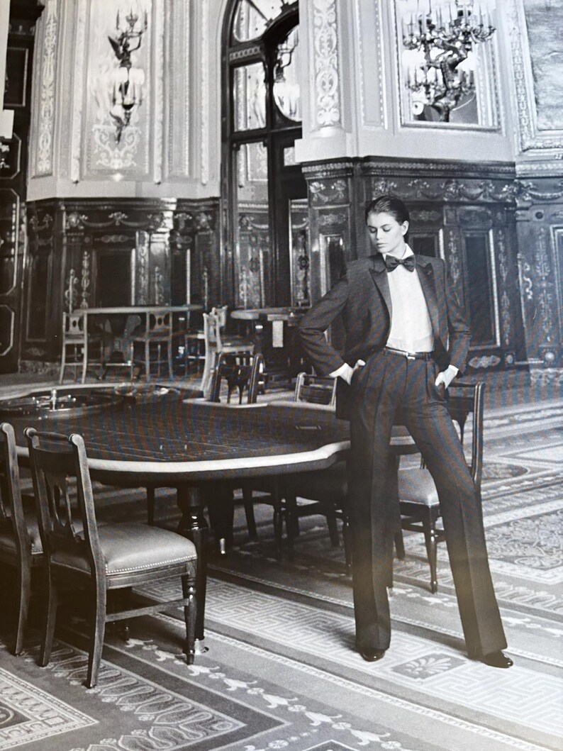 May include: A person in a dark grey suit, white shirt, and black bow tie poses in a room with a large round table and chairs. The room features ornate wall panelling and a patterned rug.