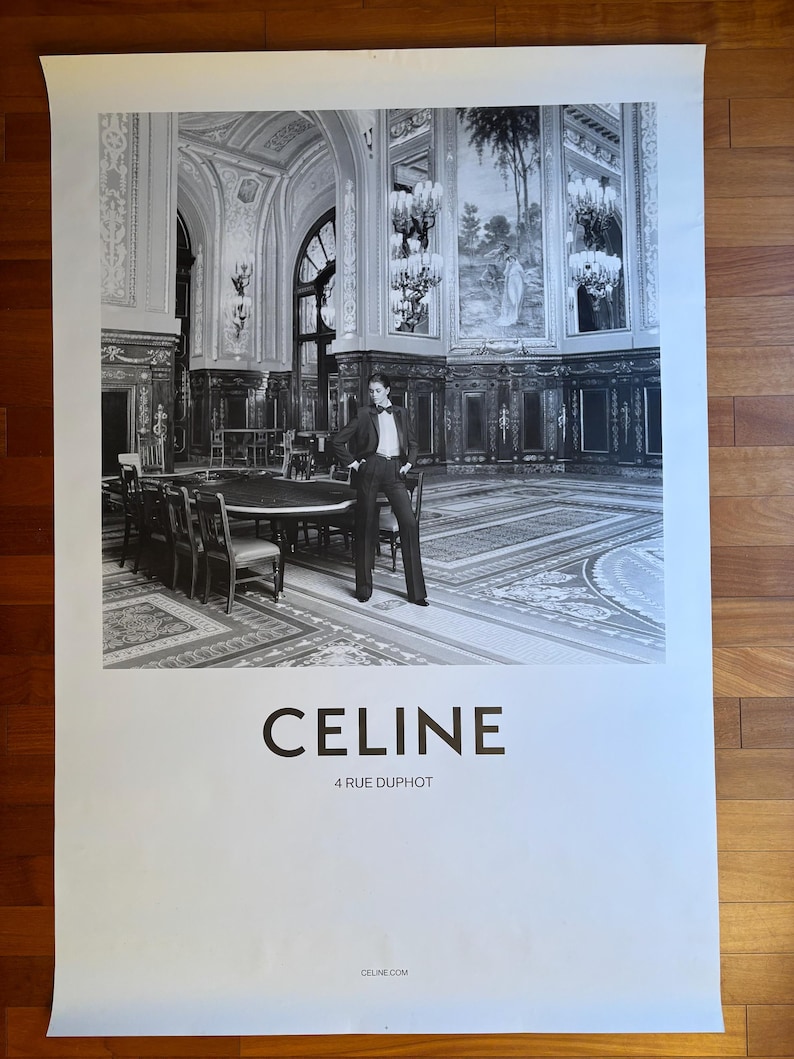May include: Monochrome poster showing a person in a suit in a lavish room with chandeliers and a large table. The word "CELINE" is printed in bold letters, with "4 RUE DUPHOT" beneath it.