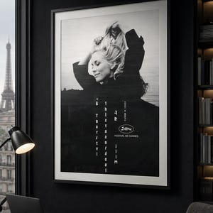 K&ouml;nnte beinhalten: Schwarz-wei&szlig; gerahmtes Poster mit einer Frau mit blonden Haaren, Text und dem Logo des Filmfestivals von Cannes. Das Poster wird in einem dunklen Raum mit einem Schreibtisch, Laptop und Blick auf den Eiffelturm gezeigt.