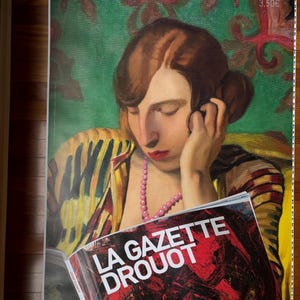May include: A woman with dark hair and red lipstick is reading a magazine called "La Gazette Drouot". The magazine cover is red with white text. The woman is wearing a yellow and black striped shirt with a pink beaded necklace.