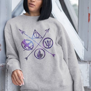 Puede incluir: Una sudadera gris con estampado de galaxia que presenta cuatro símbolos: un triángulo con un círculo dentro, un pájaro en un círculo, un tridente en un círculo y una llama en un círculo. Los símbolos están rodeados por cuatro flechas.
