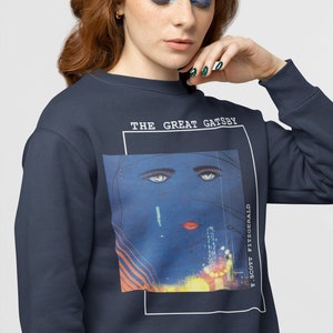 May include: A navy blue sweatshirt with a graphic print of a woman's face with blue eyes and red lips. The text "THE GREAT GATSBY" and "F. SCOTT FITZGERALD" are printed on the sweatshirt.