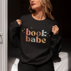 May include: Black sweatshirt with the text "book babe" in colorful retro-style lettering with stars on either side.
