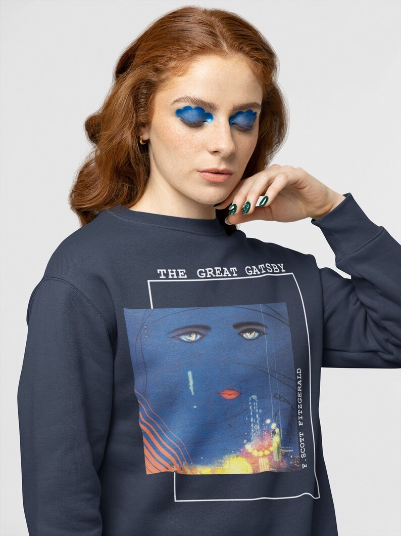 May include: A navy blue sweatshirt with a graphic print of a woman's face with blue eyes and red lips. The text "THE GREAT GATSBY" and "F. SCOTT FITZGERALD" are printed on the sweatshirt.