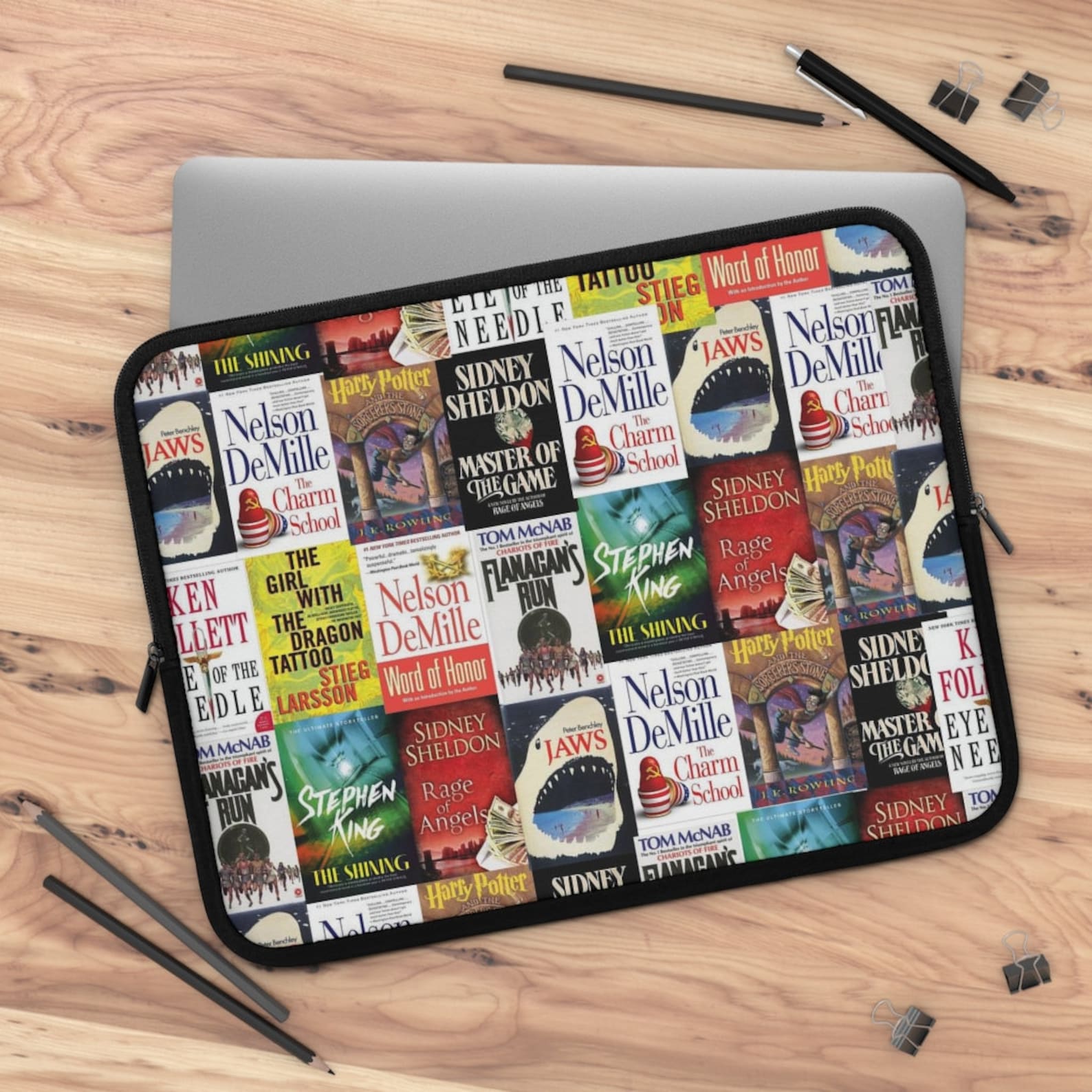 A laptop case with a collage of several different book covers sitting on a silver laptop that rests on a light wood background.