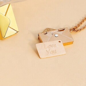 May include: Three necklaces with envelope pendants. One pendant is gold with a yellow enamel finish, and the other two are silver and gold. One gold pendant has a small tag that reads "Love You". The necklaces are on a light beige surface.