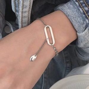 Puede incluir: Una pulsera de plata con un eslabón ovalado alargado y un dije en forma de lágrima. La pulsera está en una cadena fina con pequeños detalles de cuentas. La joya se usa en la muñeca, con un anillo de plata visible.