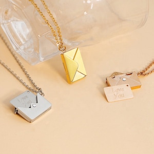 May include: Three necklaces with envelope pendants. One is silver with a square envelope that opens to reveal "Love You". Another is gold with a yellow envelope. The third is rose gold with an open envelope and a small tag that says "Love You".