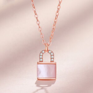 以下が含まれることがあります： ローズゴールドのネックレス。南京錠のペンダントが付いています。ペンダントは長方形で、真珠のような光沢があり、小さな透明なクリスタルで飾られた湾曲した上部があります。チェーンとペンダントは同じローズゴールドの色です。