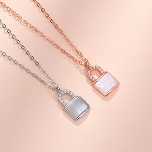 以下が含まれることがあります： 南京錠のペンダントが付いた2つのネックレス。1つはシルバー、もう1つはローズゴールドです。各ペンダントは長方形の真珠母貝の中心部と、キラキラ光るクリスタルがちりばめられたベイルが付いています。チェーンは繊細で、背景はソフトピンクです。