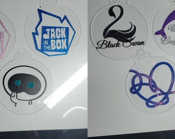 May include: Clear acrylic ornaments with various designs. One features a pink glitter design with the text "Jack in the Box". Another has a blue glitter design with the same text. Other ornaments include a black swan, a purple dolphin with the text "We are Bulletproof", and a blue and purple heart design.
