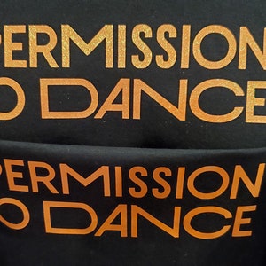 May include: Black t-shirt with the words "PERMISSION TO DANCE" in gold glitter lettering. The shirt is folded, and the text is visible on both the front and back. A pink pin is used to hold the shirt together.