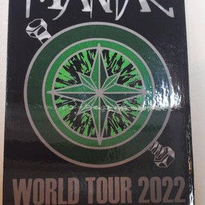 Puede incluir: Pegatina negra con la palabra "MANIAC" en blanco, sobre un diseño de brújula verde y negro. Debajo, "WORLD TOUR 2022" en plata. La pegatina es rectangular.