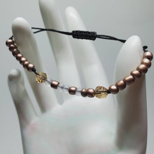 May include: A brown and white beaded bracelet with two clear crystal accents. The bracelet is made with a black cord and has a sliding knot closure.