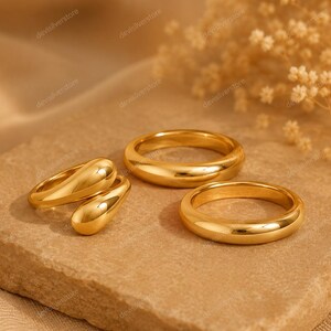 May include: A set of three gold-toned rings consisting of two smooth, plain bands and one open-style ring featuring two rounded, teardrop-shaped ends. The rings have a polished, reflective finish and are arranged together on a textured surface.