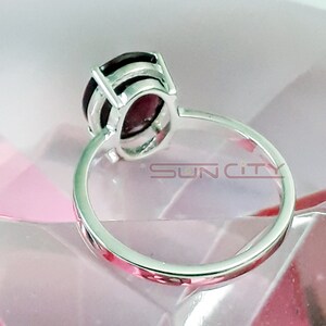 May include: A silver ring with a dark red oval gemstone set in a silver bezel. The ring is on a pink surface.