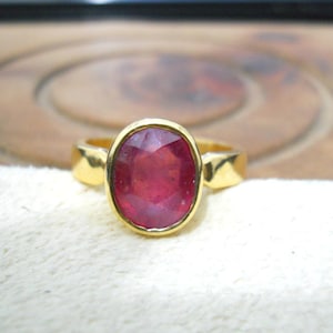 Peut inclure: Une bague en or ornée d'un grand rubis ovale. Le rubis est d'un rouge profond et est serti dans une monture en or. La bague est présentée sur une surface blanche.
