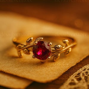 Può includere: Un anello color oro con una grande gemma rossa rotonda al centro. L'anello presenta un design a foglie sulla fascia, con piccole foglie che si estendono attorno alla gemma. L'anello è posto su una superficie strutturata e marrone.