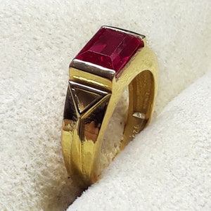 Op de afbeelding: Een gouden ring met een grote, rechthoekige, rode edelsteen. De ring heeft een driehoekig ontwerp op de band.