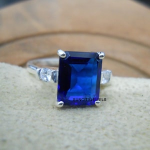 Può includere: Un anello d'argento con una grande pietra preziosa rettangolare blu scuro. Due pietre preziose chiare più piccole affiancano la pietra principale. Il testo "SUNCITYJEWELS" è visibile.
