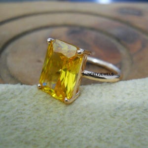Può includere: Un anello d'oro con una grande gemma gialla rettangolare. La fascia e l'incastonatura dell'anello sono dorate e la gemma è di un giallo vibrante e trasparente. L'anello è posto su uno sfondo morbido e chiaro.