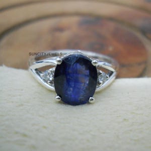 Può includere: Un anello d'argento con una grande pietra preziosa ovale blu scuro al centro. Due pietre preziose più piccole e chiare affiancano la pietra principale. L'anello è appoggiato su una superficie color crema. Il testo "SUNCITYJEWELS" è visibile.