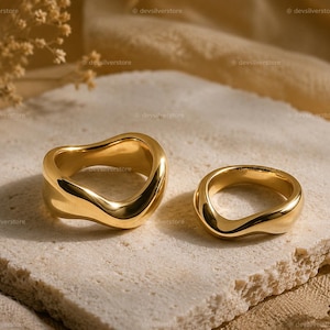 May include: Two gold-toned rings with a smooth, polished finish and an irregular, wavy, organic band shape. The rings feature a reflective surface that catches the light, showcasing a continuous, flowing design without any gemstones or engravings.