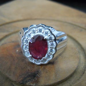 Può includere: Un anello d'argento con una grande gemma ovale rosso scuro al centro, circondata da pietre più piccole e chiare. L'anello ha una fascia a coste e un'incastonatura decorativa. L'anello è su una superficie di legno.