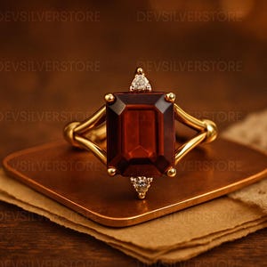 Pode incluir: Um anel de ouro com uma grande pedra preciosa retangular vermelho escuro como peça central. O anel tem um design de haste dividida e é adornado com pequenos diamantes. O anel é exibido em uma pequena superfície quadrada de cor cobre.