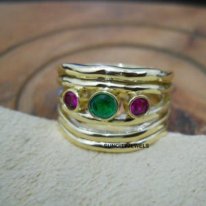 May include: A gold ring with a layered design. It features a central emerald-green gemstone, with two smaller, round, magenta gemstones. The ring has a textured, wavy band design. The text "SUNCITY JEWELS" is visible.