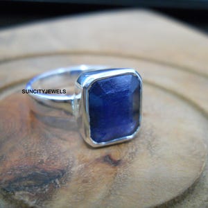 Peut inclure: Une bague en argent avec une grosse pierre précieuse rectangulaire bleu foncé. La bague est posée sur une surface en bois. Le texte "SUNCITYJEWELS" est visible sur la bague.