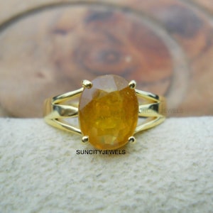 Peut inclure: Une bague en or ornée d'une grosse pierre jaune de forme ovale. La bague présente un design à tige fendue et est posée sur une surface de couleur neutre. Le texte "SUNCITYJEWELS" est visible sur la bague.