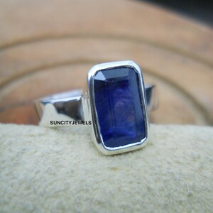 Può includere: Un anello d'argento con una grande pietra preziosa rettangolare blu scuro. La pietra preziosa è incastonata in una lunetta. L'anello è esposto su una superficie di colore chiaro. Il testo "SUNCITYJEWELS" è visibile.
