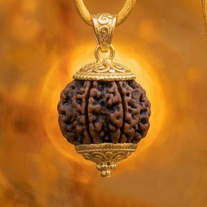 Peut inclure: Un pendentif composé d'une perle rudraksha sphérique et texturée brun foncé, enserrée entre deux capuchons en métal doré ornés. Le pendentif présente une bélière décorative dorée sur le dessus et est suspendu à un cordon doré assorti.