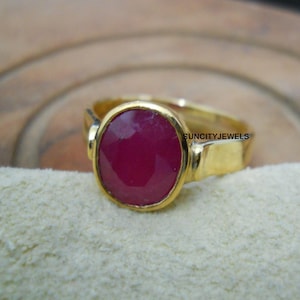May include: A gold ring featuring a large, oval-cut ruby gemstone. The ruby is a deep, rich red color, set in a gold band. The ring has a simple, elegant design. The text "SUNCITYJEWELS" is visible on the ring.
