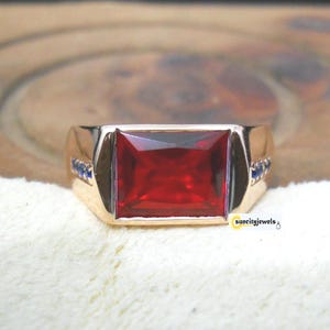 May include: A gold ring featuring a large, rectangular red gemstone. Small blue gemstones are set on either side of the main stone. The ring is presented on a white surface.