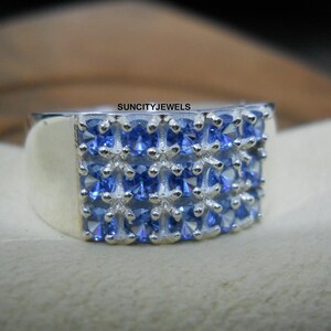 Può includere: Un anello d'argento con una superficie rettangolare, adornato con file di piccole pietre preziose blu a taglio quadrato. L'anello ha una finitura lucida e il testo "SUNCITYJEWELS" è visibile.
