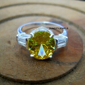 Può includere: Un anello d'argento con una grande pietra preziosa gialla a taglio ovale come elemento centrale. Due pietre preziose rettangolari più piccole affiancano la pietra principale. L'anello è posto su una superficie di legno, con il testo "SUNCITYJEWELS".